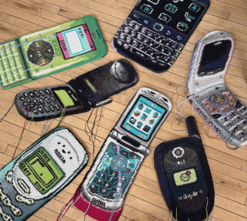 Fiber art pieces by Nicole Nikolich of dated flip phones, laid out on a hardwood floor